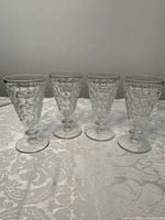 Four vintage Jeannette pressed glass water goblets arranged side by side on a patterned tablecloth.