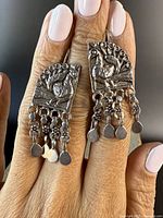 Front view of two silver earrings with peacock design and dangling parts, worn on fingers for scale.