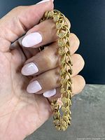 Hand holding the gold-tone braided bracelet showing chain links with rounded elements and textured floral clusters.