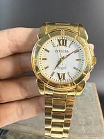 Close-up front view of Invicta Angel watch showing white face with gold Roman numerals and hands, and metal gold tone bracelet.