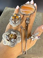 Six antique Scottish sterling silver jewelry pieces displayed on hand, showing oval agate brooch, thistle motif brooches with citrine stones, kilt pin with thistle head, and pearl-accented brooch.