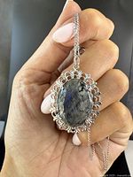 Hand holds the pendant necklace showing the intricate sterling silver filigree setting around the polished oval labradorite stone with visible blue sheen.