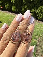 Pair of mixed metal circular orca earrings displayed on fingers outdoors showing gold-tone, copper, and silver colors with intricate orca design.