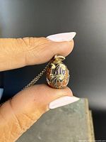 Close-up photo of the egg-shaped pendant on the sterling silver chain held between fingers, showing detailed enamel decoration with gold overlay.