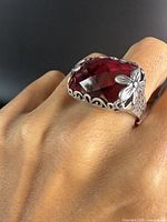 Ring on finger showing the large rectangular pink faceted stone with silver floral bezel and band on a hand with black background.