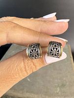 Close-up photo showing the pair of sterling silver stud earrings with gold accents and floral scrollwork design held between fingers.