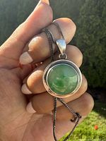 Close-up of the circular green jade pendant set in sterling silver, held between fingers showing texture and design details.