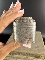 Front view showing engraved floral scroll design and hinged domed lid held between fingers for scale.