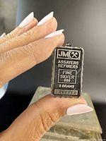 Front view of vintage rectangular pendant shaped like a silver ingot bar with J&M Assayers Refiners and silver purity markings. Held between fingers for scale.