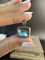 Front view showing blue-green iridescent labradorite stone with silver bezel and decorative bail held between fingers.