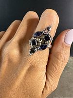 Photo of ring worn on hand showing the front cluster of natural gemstones in blue and green hues and ornate sterling silver work.