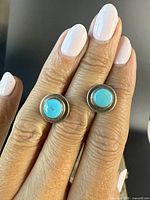 Close-up front view of the turquoise and silver stud earrings worn on fingers to show size and detail.