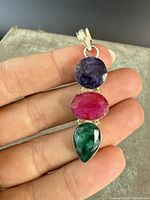 Front view of the pendant showing three faceted stones: sapphire, ruby, emerald in sterling silver bezel setting held by hand.