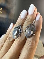 Close-up frontal view of both earrings held between fingers, showing moonstone cabochons and decorative silver work on one side.