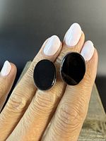 Pair of oval black onyx stud earrings shown front view on fingers to display size and design.