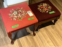 Two antique footstools side by side, salmon-pink and dark reddish-brown needlepoint upholstery with floral needlepoint designs on top, wood legs visible.