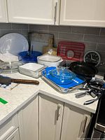 Corner kitchen countertop with assorted kitchenware including CorningWare and Pyrex bakeware, plates, utensils, clock, and red kitchen rack