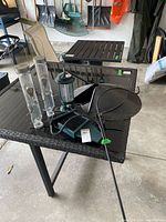 Black metal bird feeder hanger stand with curved top, two clear tube feeders, a green metal lantern-style feeder, black shallow tray/dish, and green pointed rod accessory laid on a black glass-top table.