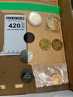 Top view of assorted collectible coins and tokens displayed on a cardboard surface with a MaxSold note for scale and measurement.