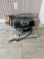 Sony boombox, CDs, VHS tape, cables, and Zenith DVD player arranged on floor against wall.