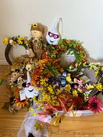 Fall decorative wreaths, floral picks, and scarecrow plush figures arranged on floor against wall.
