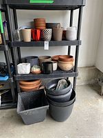 Flower pots of various materials and colors including terracotta, plastic, ceramic, glass, arranged stacked on black shelves.