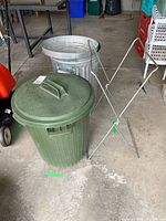 Side by side view of green plastic garbage can, galvanized metal garbage can, and foldable metal stand placed on a concrete floor in garage setting.