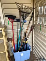 Wide view of garden tools organized in blue plastic bin inside a shed, including rakes, brooms, axe, pruning shears, shovel, and hoe