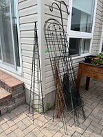 View of multiple metal garden trellises and two plant hangers leaning against exterior siding near a window.