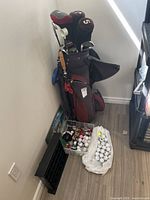 Photo shows a maroon and black golf bag standing upright containing multiple golf clubs with covers. Next to the bag are two containers filled with numerous golf balls and various golf accessories including tees and gloves.