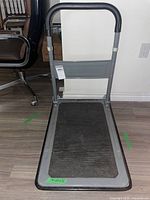 Front view of gray metal platform hand truck with broad curved handle and black rubber mat on platform.