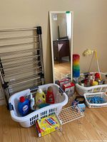 Full view of lot items: metal drying rack, two white laundry baskets with cleaning liquids, white basket with clothespins, tall floor mirror, colorful dusters, and box of SOS pads.