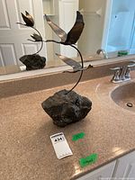Full view of the metal sculpture with two birds flying above a stone base, showing overall height and display setting on a bathroom counter in front of a mirror.