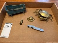 Overview of all metal items in the lot inside a cardboard box, showing a small blue-green metal cradle, brass crab, brass owl, corkscrew, and a small brass knob or button.
