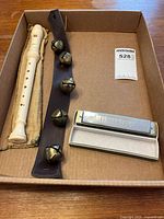 Photo of all items in a cardboard tray: cream plastic recorder in pouch, leather strap sleigh bells with four large bells, and silver Mississippi harmonica in open plastic case.