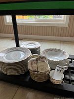 Full display of the china set piled on a table to show quantity and pattern. Includes dinner plates, dessert plates, bowls, cups, and saucers with pink floral design on white porcelain.