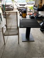 Photo shows the outdoor patio chair with beige mesh back and seat alongside the black square patio table with pedestal base in garage environment.