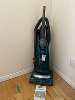Full front view of the upright vacuum cleaner standing on wooden floor in corner near electrical outlet, manual placed in front.
