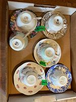 Overview of all 8 cups and saucers from Shelley and Paragon in a cardboard box, showing different floral and gold pattern designs.