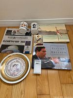 Photo of various royal family memorabilia items arranged on floor including a commemorative plate, two mugs, newspapers, postcards, and books.