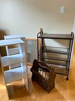 Photo showing two white shelving units, dark wooden magazine rack, black metal and wood utility cart, and framed mirror against wall.