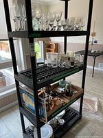 Full view of the tall black plastic shelving unit with the assortment of glassware, decorative items in a box, and plates on the bottom shelf.