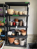 Front view of the four-tier black plastic shelving unit filled with various flower pots to illustrate shelf size, mainly for reference showing the shelf itself is for sale only.