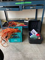 Lot contents showing multiple extension cords, a green crate with items, rod-shaped tools, and a black plastic box on floor below a workbench.