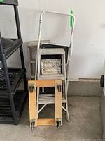 Photo showing three metal step stools stacked behind a wooden dolly with caster wheels.