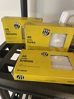 Display of yellow boxes containing 48 plastic spoons, forks, and knives each, No Name brand.
