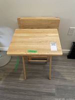 Three wooden folding TV tables stacked and folded against a wall on hardwood floor.