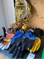 Eight pairs of work gloves in various colors and sizes, coiled ropes, two yellow flashlights, and bungee cords on a workbench against a pegboard.