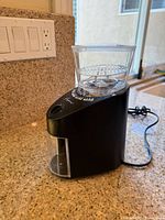 Side view of the Capresso Infinity burr grinder on a kitchen counter showing its black body, clear plastic hopper, and power cord.