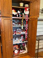 Wooden cabinet with upper shelves showing various plush Santa and snowman dolls, ceramic figures, mugs, and hanging felt candy cane and heart garlands.